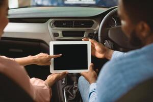 Afro Couple Using Digital Tablet With Empty Screen Sitting In New Car In Dealership Center. Mockup, Back View, Selective Focus photo
