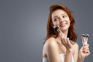 Red-Haired Young Female Holding Cosmetic Brushes Near Face Making Makeup Posing Looking Aside In Studio On Gray Background. Make-Up Artists Concept. Panorama, Blank Space photo