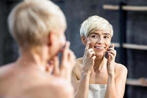 Happy Middle-Aged Woman Applying Facial Cream Moisturizer On Face Standing Wrapped In Towel In Bathroom. Mature Skin Treatment. Selective Focus photo