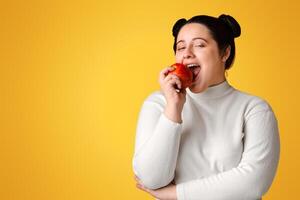 Positive Young Plus Size Girl Eating Red Apple, Posing Over Yellow Background In Studio And Looking At Camera, Empty Space photo