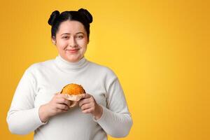 Positive Oversize Girl Holding Tasty Hamburger And Smiling At Camera, Posing On Yellow Background With Free Space photo