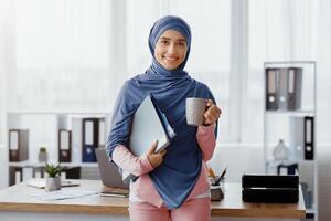 Trainee in office concept. Portrait of smiling young woman in hijab with coffee cup and document folders standing near desk photo