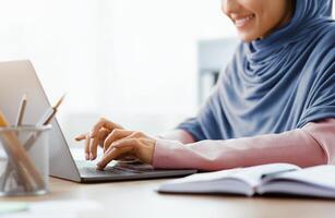 Unrecognizable muslim woman in hijab working on laptop computer in office, typing on keyboard and smiling, cropped image with empty space photo