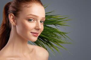 Ginger-Haired Female Posing In Front Of Exotic Green Plant Smiling To Camera In Studio Over Gray Background. Panorama With Empty Space For Natural Cosmetics Advert photo