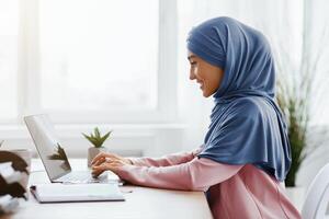 Office Work For Islamic Women. Happy Muslim Girl In Hijab Working On Laptop Computer At Desk, Typing On Keyboard, Side View With Free Space photo