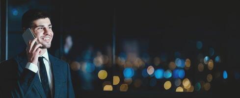 Smiling Entrepreneur Talking On Phone Standing Near Window With City View In Modern Office At Night. Business Communication. Copy Space photo