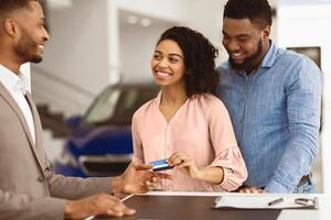 Buying Car. African American Spouses Giving Credit Card To Salesman Standing In Automobile Dealership Office. Selective Focus photo