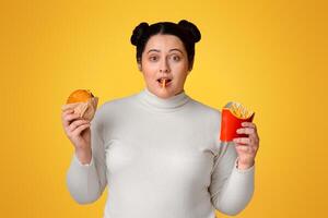 Overweight young woman with french fries serving and burger in hands, enjoying eating fast foods, standing over yellow background, copy space photo