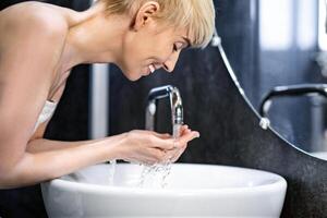 Joyful Mature Woman Washing Face Doing Morning Beauty Routine Standing Wrapped In Towel In Bathroom. Selective Focus photo