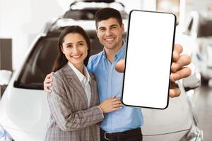 Ownership Concept. Young family embracing and standing together in front of auto in showroom, showing car keys at camera photo