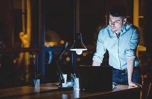 Young Businessman Working On Laptop Standing In Modern Office Late At Night. Business Lifestyle. Copy Space photo