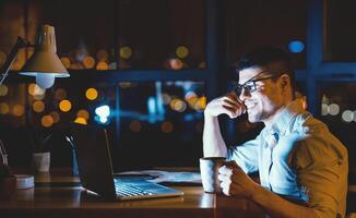 Business Guy Sitting At Laptop Having Coffee In Modern Office At Night. Corporate Lifestyle. Copy Space photo