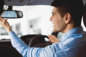 Close up back view portrait of smiling man in shirt adjusting rearview mirror while sitting in his car, selective focus photo