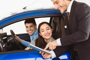 End Of A Deal. Car seller showing digital tablet to young couple who sitting in auto at dealership office photo