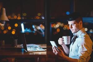 Business App. Businessman Using Smartphone Texting In Corporate Chat Working On Laptop Overnight In Office. Empty Space photo