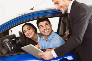 Check Out The Characteristics. Happy couple sitting in salon of new car, vendor showing tablet to them photo