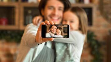 Happy dad and daughter taking selfie on smartphone while cooking at kitchen, little girl and her father hugging and smiling at camera, selective focus photo