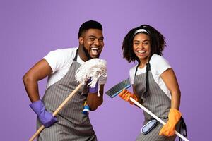 Positive african couple fooling together while making spring cleaning, singing into mop and having fun over pink background with copy space photo