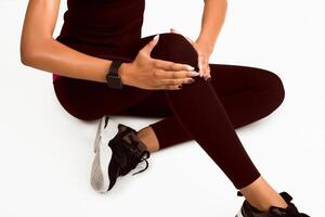 Unrecognizable Black Woman Massaging Knee Suffering From Pain Sitting On White Studio Background. Fitness And Bad Joints. Cropped photo