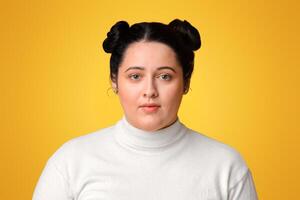 Portrait of young plus size girl with double bun hairstyle standing over yellow background and looking at camera, free space photo