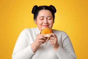Plump Hungry Girl Holding Burger In Hands And Sniffing It With Pleased Face Expression, Standing Over Yellow Background With Copy Space photo