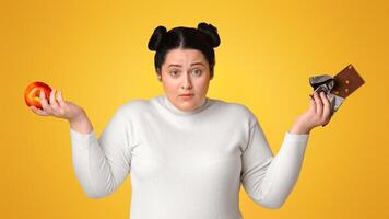 Puzzled Overweight Woman Holding Chocolate Bar And Apple In Hands, Can't Decide What To Choose. Yellow Studio Background, Panorama photo