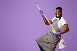 Portrait Of Funny Black Man In Apron With Mop In Hands Posing Over Pink Studio Background, Using Handle As Guitar, Fooling With Household Tool photo