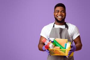 Portrait of positive black man holding basket with detergents and cleaning tools, isolated on pink background with copy space photo