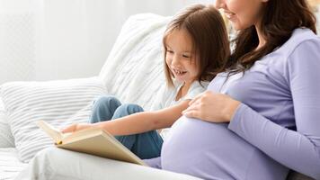 Little daughter and pregnant mom enjoying time together, sitting on sofa at home and reading book, empty space photo
