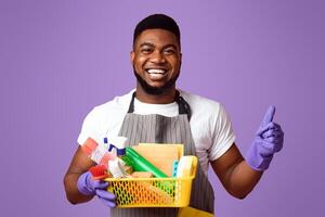 Cleaning With Pleasure. Positive Black Man Holding Container With Detergents And Household Supplies And Showing Thumb Up Over Pink Background photo