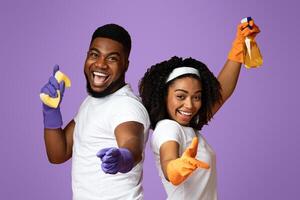 Housekeeping and cleaning service concept. Joyful black couple holding spray detergent and sponge and pointing fingers at camera, pink background photo