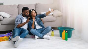 Happy Black Couple Taking Selfie On Smartphone After Tidying Flat Together, Relaxing On Floor After Spring-Cleaning, Showing Peace Gestures photo