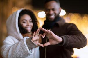 I Love You. Closeup of cheerful positive african american couple making heart shape with fingers walking together in the evening, looking at hands, selective focus, blurred background. Valentine's day photo