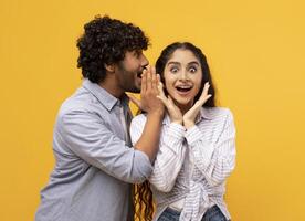Shock, gossip, share advice. Young indian man whispering to lady on ear, happy woman with open mouth excited and surprised, posing on yellow studio background photo