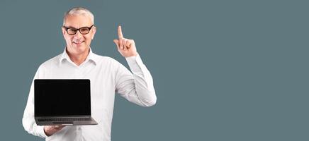 Useful Website. Middle-aged man pointing up and holding laptop with blank black screen over blue studio wall, mock up photo