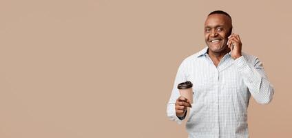 Pleasant talk. Happy mature african american man with coffee talking on mobile phone, orange studio background photo