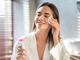 hermosa sonriente mujer limpieza piel con micelar agua y algodón almohadilla mientras haciendo diario belleza rutina en baño, atractivo joven hembra disfrutando hogar protección de la piel tratos, Copiar espacio foto