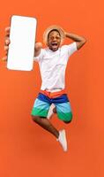 Cool mobile offer. Excited jumping black guy demonstrating smartphone with white screen on orange studio background, collage with mockup copy space for app or website design photo