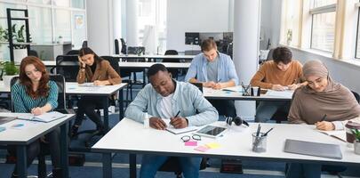 Modern Education Concept. Portrait of diverse group of students sitting at desks in classroom at university, taking test, entrance examination or writing notes in notebook, selective focus photo