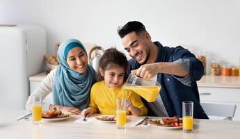 Portrait Of Happy Middle Eastern Family Of Three Having Breakfast In Kitchen Together, Arab Parents With Cute Little Daughter Enjoying Tasty Food And Drinking Orange Juice While Sitting At Table photo