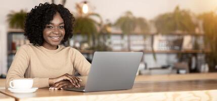 Cheerful afro girl smiling at camera while working online at cafe, using laptop, drinking coffee, copy space photo