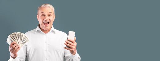 Making Money Via Internet. Excited mature man showing bunch of money, holding smartphone, free space, blue studio wall photo