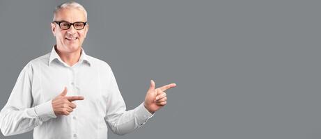 Cool Offer. Smiling mature man in glasses pointing two index fingers aside at copyspace, gray studio wall, panorama photo