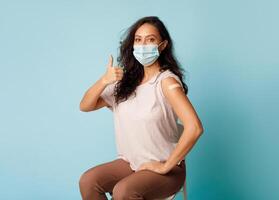 Covid-19 Vaccination. Vaccinated Woman Gesturing Thumbs Up Showing Arm With Plaster Bandage After Coronavirus Vaccine Injection On Blue Background, Wearing Protective Face Mask. Antivital Immunization photo