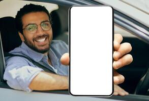 Closeup of young smiling Indian man in glasses showing big mobile phone with white empty screen for mock up sitting on driver seat in new luxury car, holding gadget in hand, selective focus on device photo