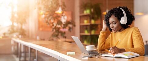 Smiling black girl with wireless headset studying online, using laptop at cafe, taking notes, copy space photo