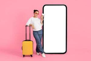 Asian young woman stands beside a vibrant yellow suitcase, holding a smartphone and posing confidently against a large blank screen in a studio setting with a pink background. photo
