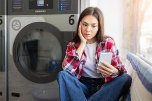 A young woman is sitting cross-legged on the floor in a laundry room, looking at her phone with a thoughtful expression. Clothes are being washed in the background. photo