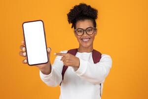 A cheerful young woman with curly hair holds a smartphone in front of her, pointing at the blank screen. The bright orange background adds energy to the scene, creating a lively atmosphere. photo