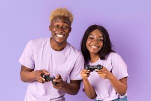 Cheerful African American couple with joysticks playing games on lilac background photo